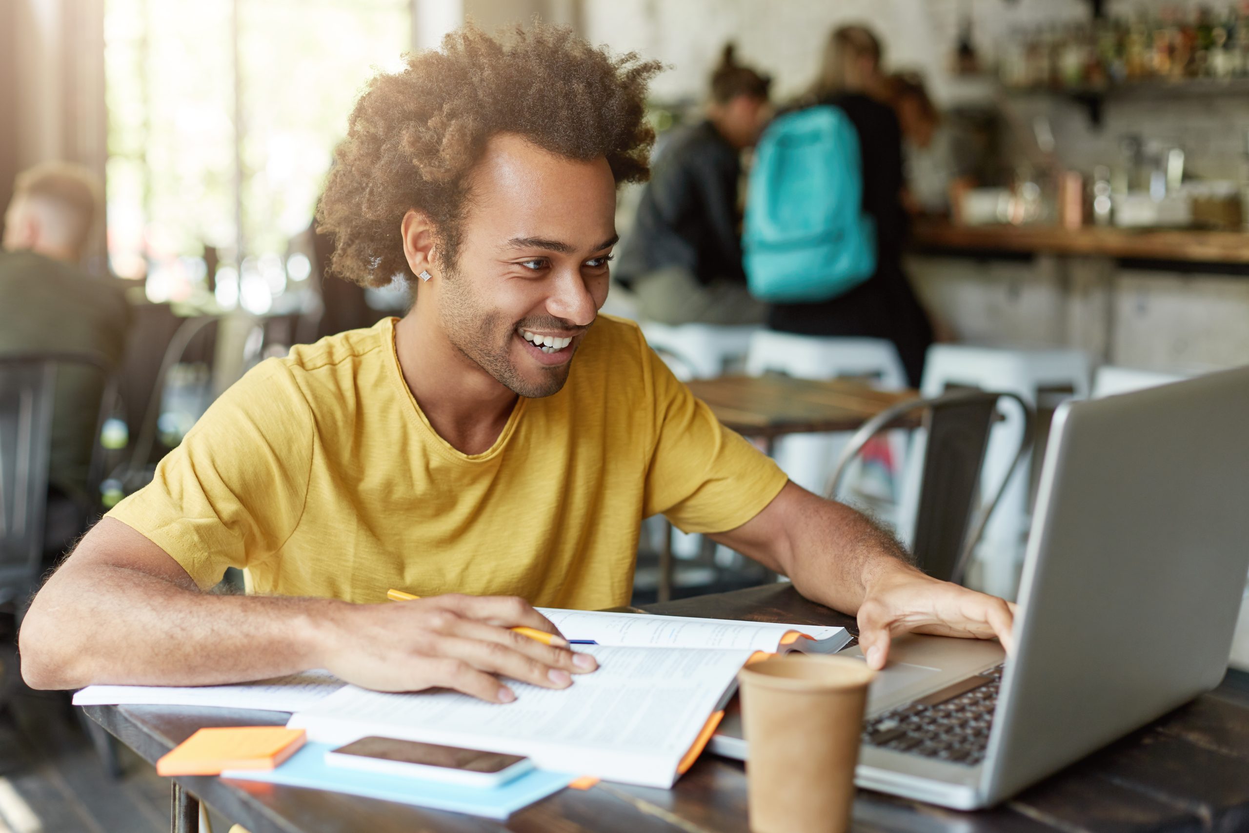 foto-interna-de-um-estudante-feliz-sexo-masculino-com-cabelo-encaracolado-vestido-casualmente-sentado-na-cafeteria-trabalhando-com-tecnologias-modernas-enquanto-estuda-olhando-com-um-sorriso-no-caderno