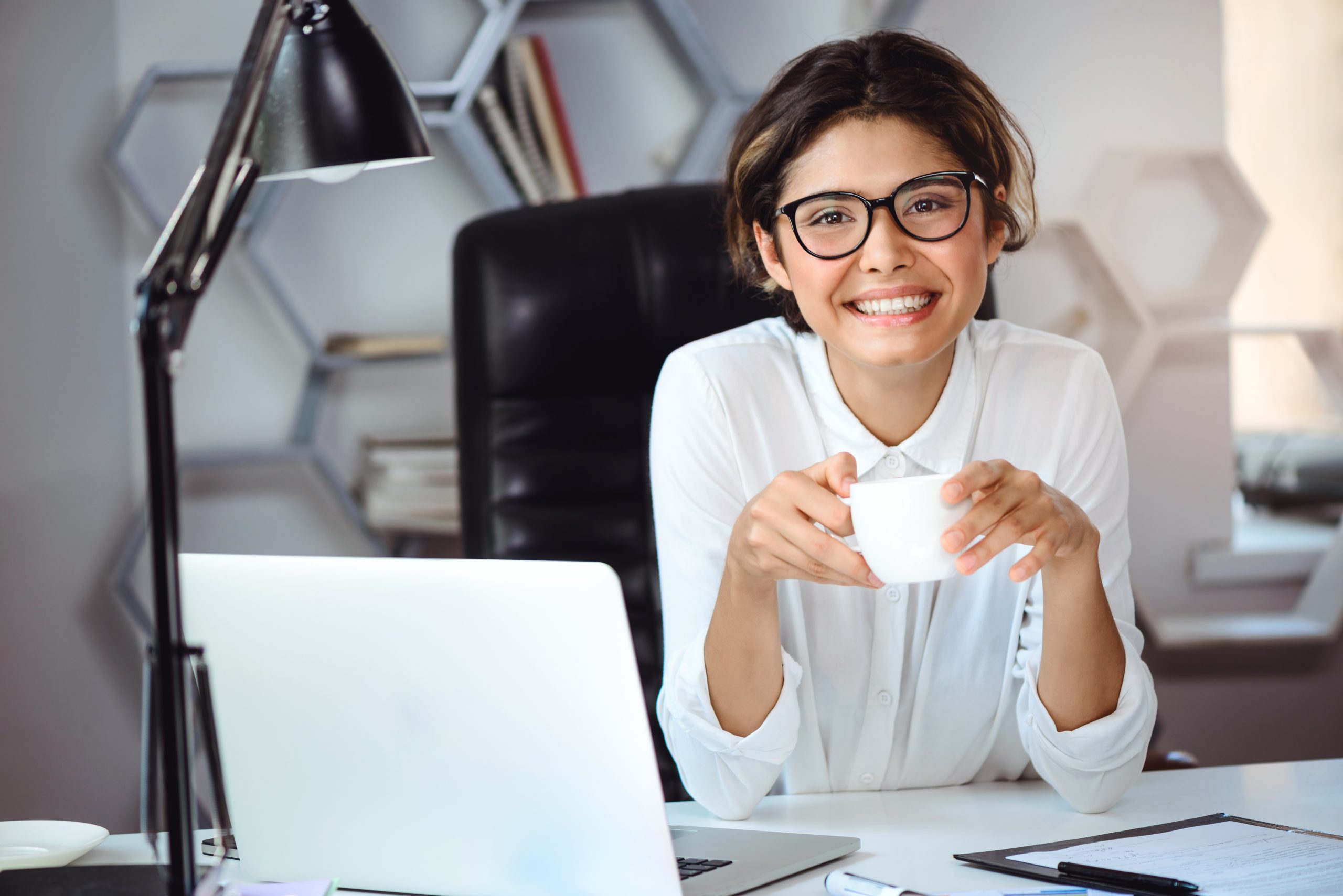 Mulher branca em um escritório com sorriso e uma xícara branca nas mãos com trajes sociais, tendo uma mesa à frente com um notebook a seu lado direito.
