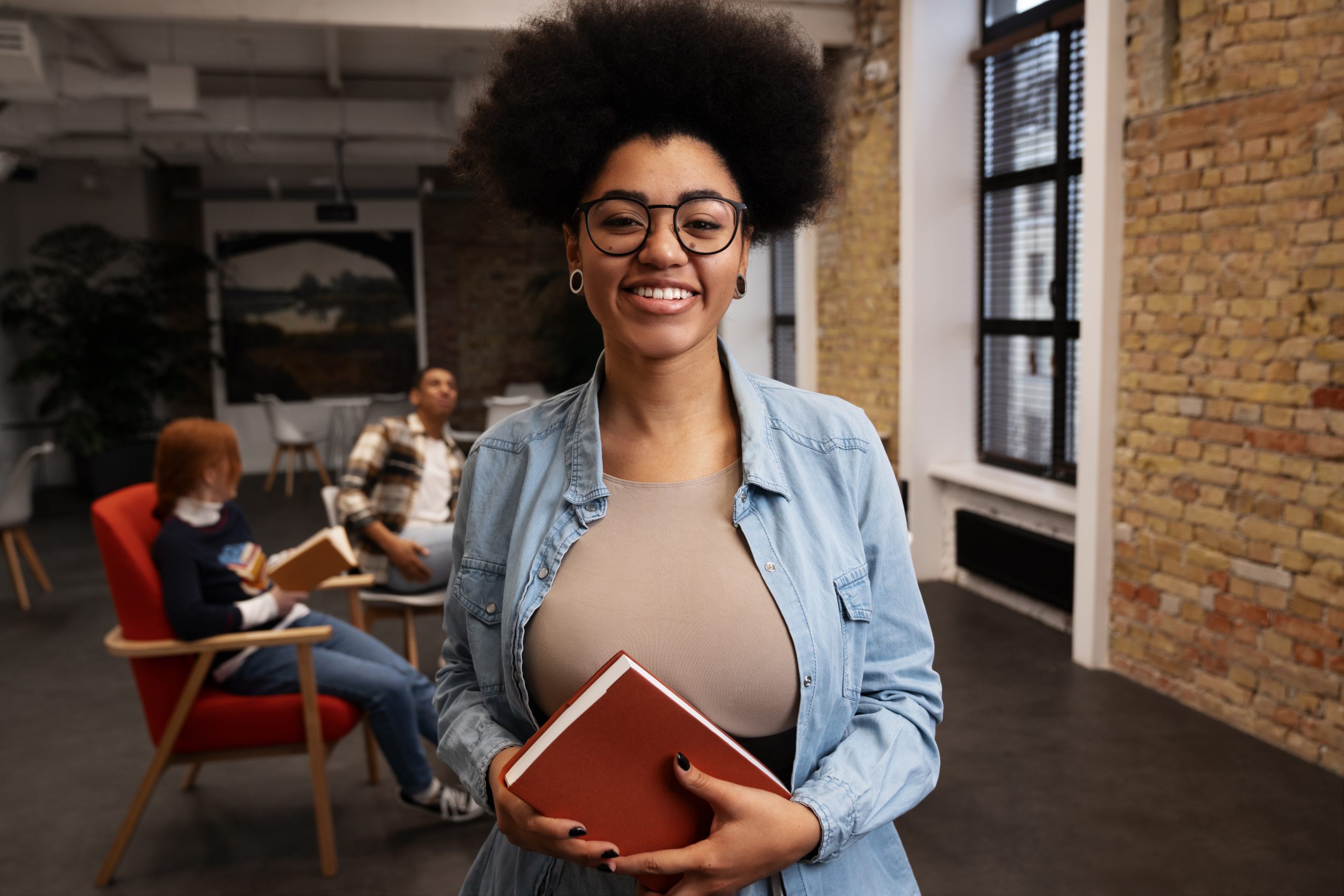mulher-negra-com-livro-na-mao-e-atras-duas-pessoas-conversando-em-biblioteca