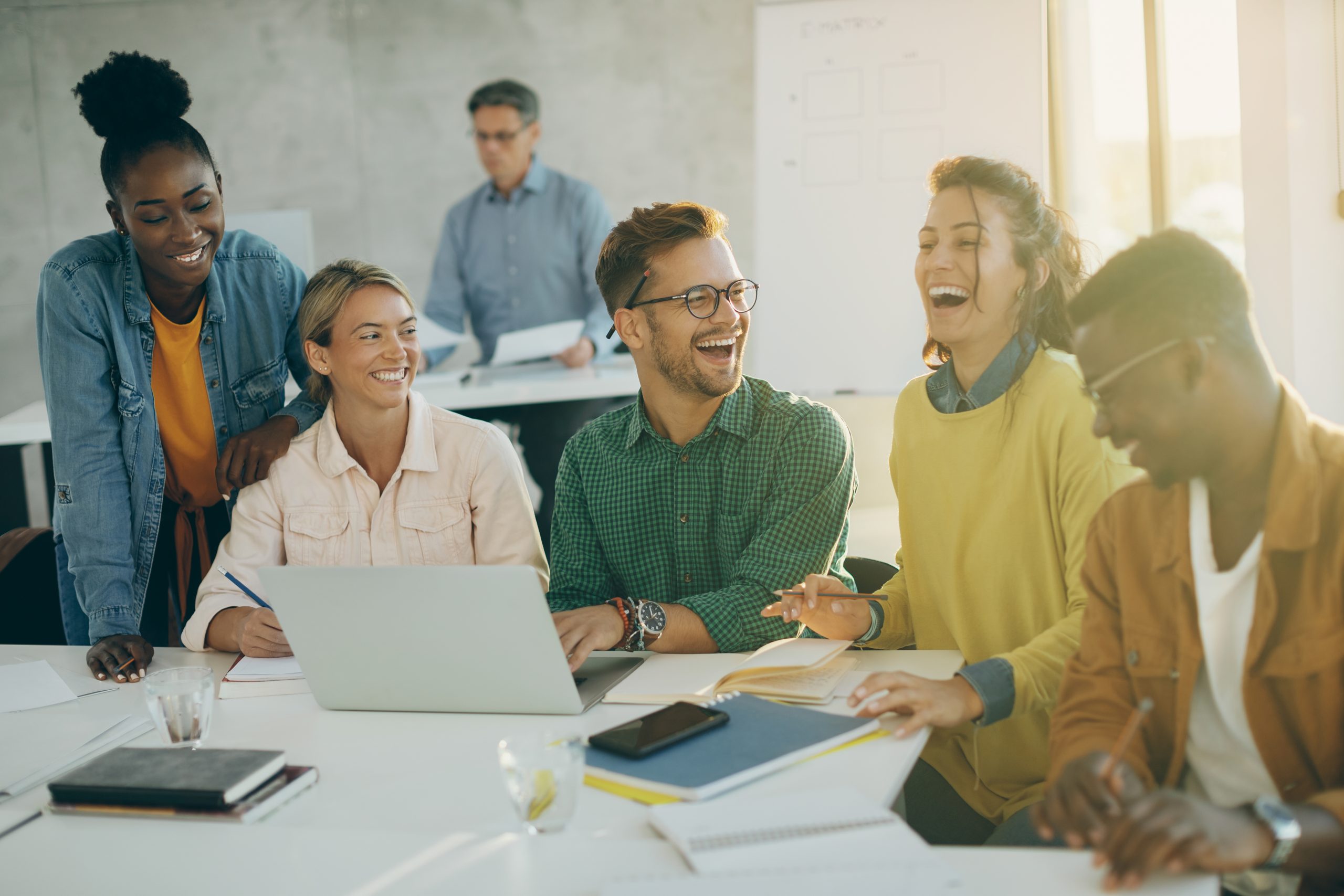 grupo-de-estudantes-felizes-se-divertindo-e-rindo-enquanto-usa-o-laptop-e-estuda-na-sala-de-aula-o-professor-deles-esta-em-segundo-plano