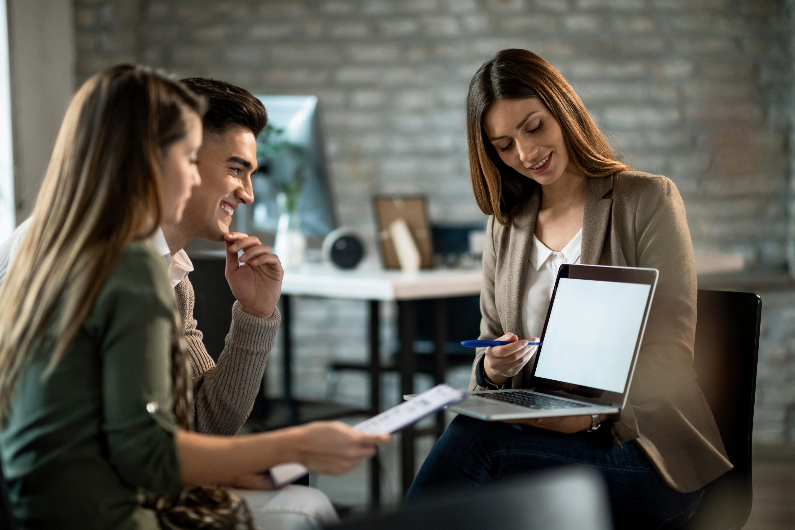 agente-imobiliario-sorridente-usando-um-computador-enquanto-se-encontra-com-um-jovem-casal-e-faz-planos-de-investimento