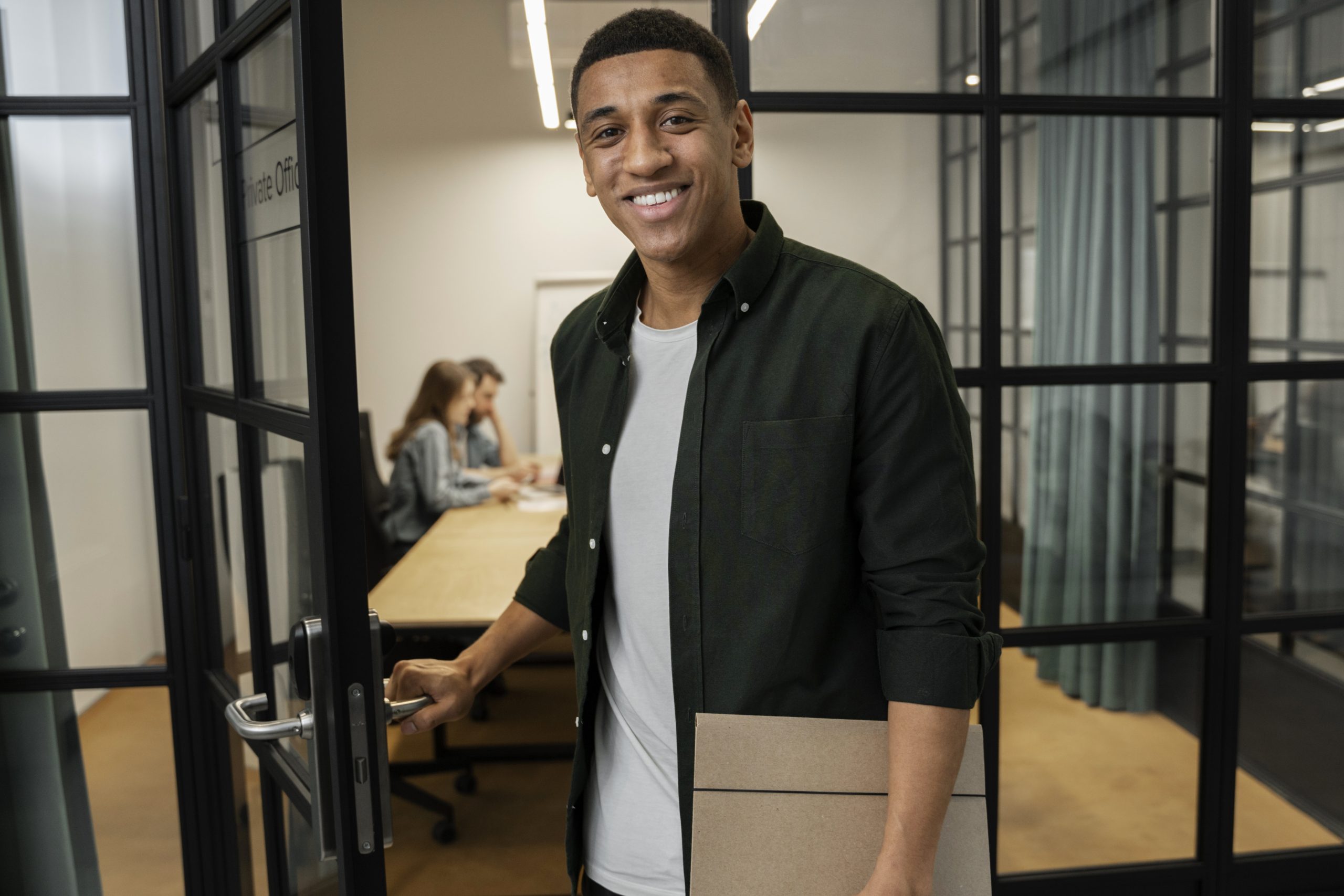 Em um escritório, vemos um homem negro encarando a câmera com um sorriso , tendo uma pasta em sua mão direita e a maçaneta de uma porta de vidro aberta em sua mão esquerda, ele parece sair do ambiente corporativo.