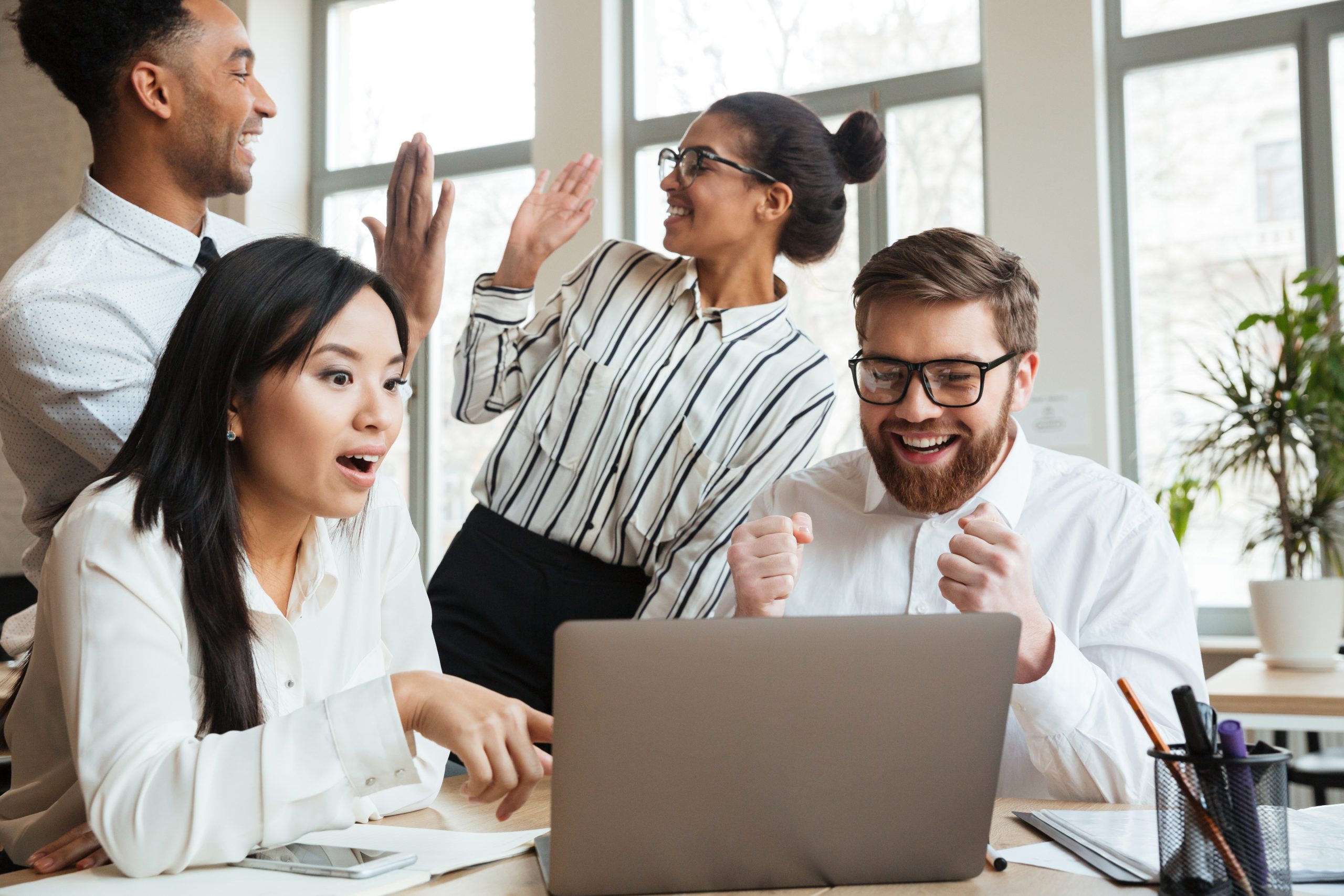 Quatro colegas de trabalho em um escritório, sendo um homem em pé negro ao fundo, perto de uma mulher branca de pé, tendo à frente uma mulher oriental sentada e um homem de óculos branco sentado, ambos olhando para um notebook. Os dois de trás comemoram e os dois da frente estão com expressões felizes e surpresas ao olhar o notebook.
